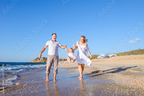 Happy family of three - pregnant mother, father and daughter holding hands and having fun walking on the beach. Family vacation, travel concept. Bright sunlight. Copy space.