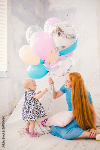 Children's holiday theme. Young beautiful mother gives, gives to the child a bunch of balloons sitting on the floor of the house