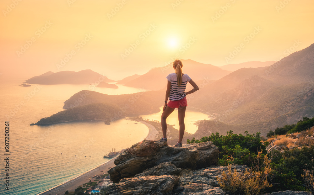 Young woman standing on the top of rock and looking at the seashore and mountains at colorful sunset in summer. Landscape with girl, sea, mountain ridges and orange sunlight. Travel. Oludeniz, Turkey