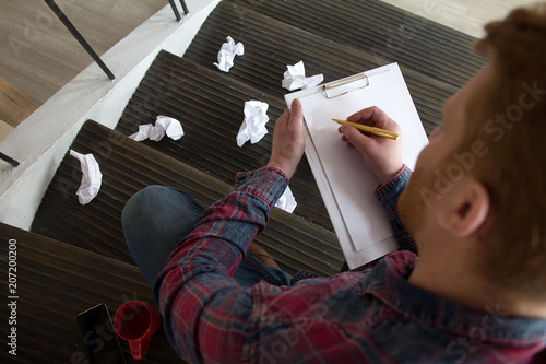 Ginger man taking notes on sheet of white paper. Guy writing on A4 paper on clip note pad while sitting on staircase with crampled paper balls around him.