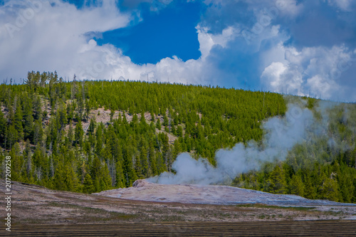 Beautiful view of old faithful Geyser Basin located in Yellowstone National Park, surrounded by vapor with a green mountain behind in gorgeous sunny day photo