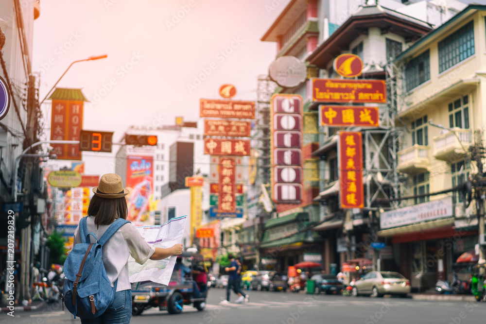 Naklejka premium Młody azjatycki podróżnik kobieta z niebieskim plecakiem i kapeluszem hipster patrząc mapę na drodze z tłem Tajlandii tuk tuk w China Town Bangkok. Podróżowanie po Bangkoku w Tajlandii