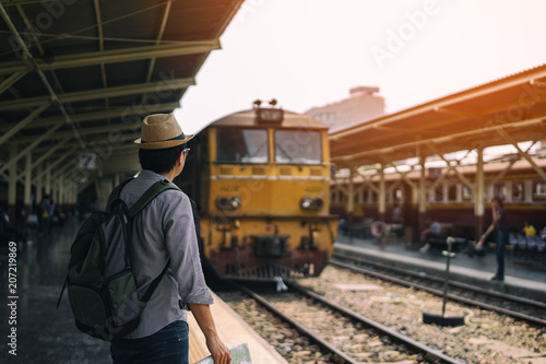 Young asian man traveler with backpack and hat standing and holding map waiting train for travel at train station in Bangkok city Thailand. Traveling in Bangkok Thailand