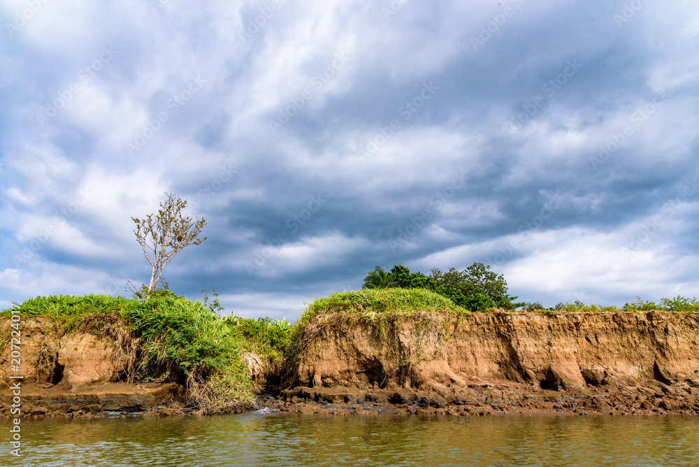 Erosion in a stormy landscape on the bank of the Tarcoles River, Costa Rica, side stream, tree and bushes on the river bank, and a stormy sky in the background
