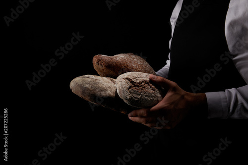 cropped image of male baker in apron holding various types of bread isolated on black background