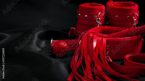Bondage, kinky adult sex games, kink and BDSM lifestyle concept with a pair of red leather handcuffs, flogger, ball gag and a coller with a leash attached on black silk with copy space