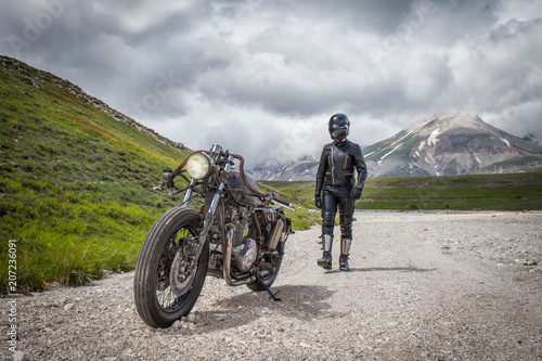 Biker with black leather clothes  walking near his custom rat motorcycle in a desolated mountain land. Post apocalyptic concept