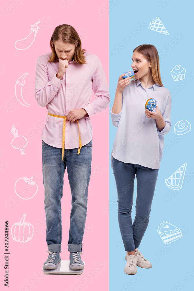 Extra weight. Emotional worried man looking at the scales while standing on them with a laughing girlfriend by his side Extra weight. Emotional worried man looking at the scales while standing on them with a laughing girlfriend by his side