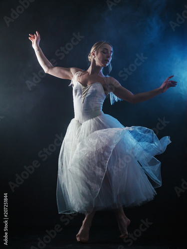 Ballerina is practicing her moves in dark studio. Young girl dancing with air white dress tutu, spinning around and smiling. Gracefulness and tenderness in every movement