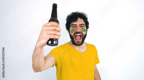 Sport fan holding a beer screaming for the triumph of his team. Man with the flag of Brazil makeup on his face and yellow t-shirt. 