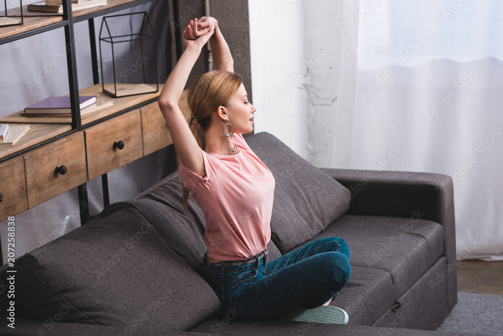 side view of attractive young woman with arms up sitting on sofa at home side view of attractive young woman with arms up sitting on sofa at home