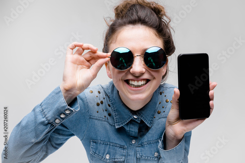 Young brunette girl in round glasses. Hairs are gathered in a bun. Girl with a black phone. © Ivan Traimak
