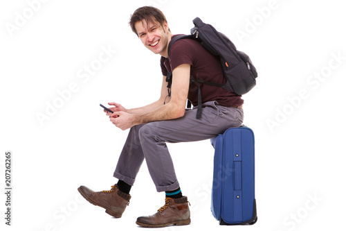 Full length happy young traveler holding cellphone and sitting on suitcase