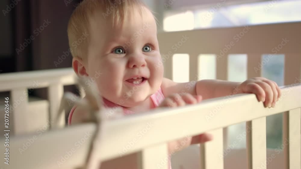 Joyful kid standing in crib. Smiling toddler standing in bed at home. Cute baby turning head in cot. Cheerful child girl playful smile. Sweet infant looking parent from bed. Child time concept Joyful kid standing in crib. Smiling toddler standing in bed at home. Cute baby turning head in cot. Cheerful child girl playful smile. Sweet infant looking parent from bed. Child time concept
