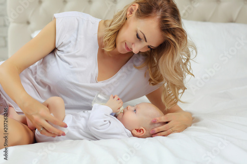 Mom feeds the baby in bed from a bottle in white clothes.