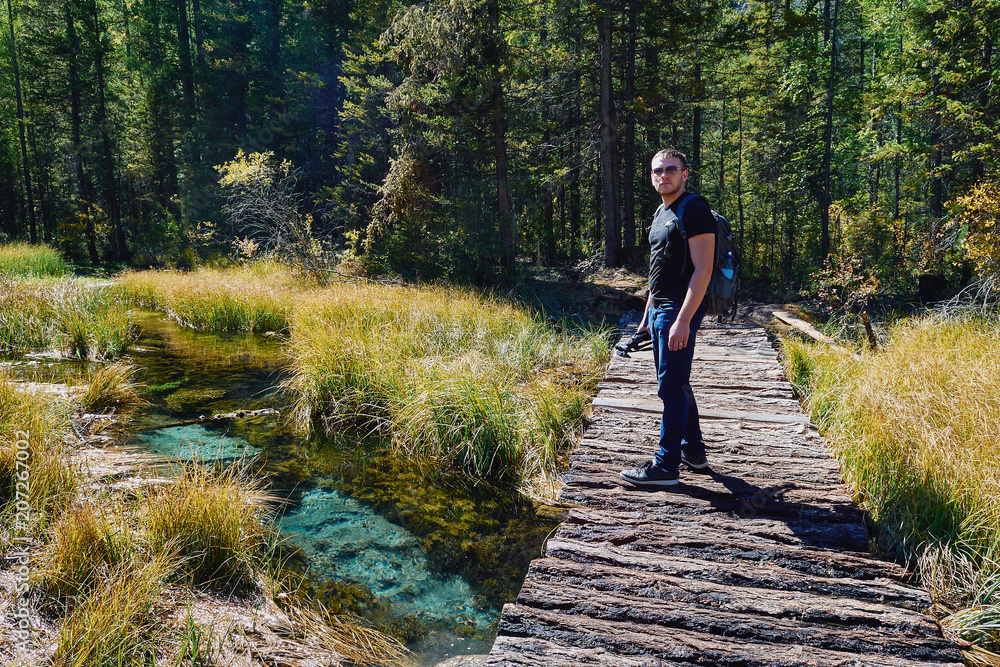 Young man with a backpack and tripod is walking along the bridge across a mountain stream into the depths of the forest towards adventures
