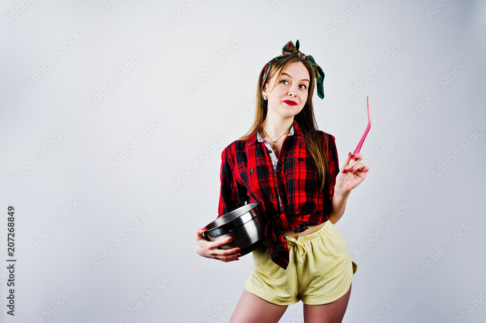 Obraz premium Young funny housewife in checkered shirt and yellow shorts pin up style with saucepan and kitchen spoon isolated on white background.
