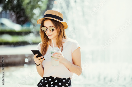 Young beauty woman with phone chating or surf in internet drinks mojito fruit cocktail against fountain in the street photo