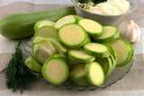 Shredded zucchini round slices on a glass plate, next to a garlic head, a whole zucchini fruit, green dill and a plate with sour cream and chopped garlic, for cooking a snack recipe.