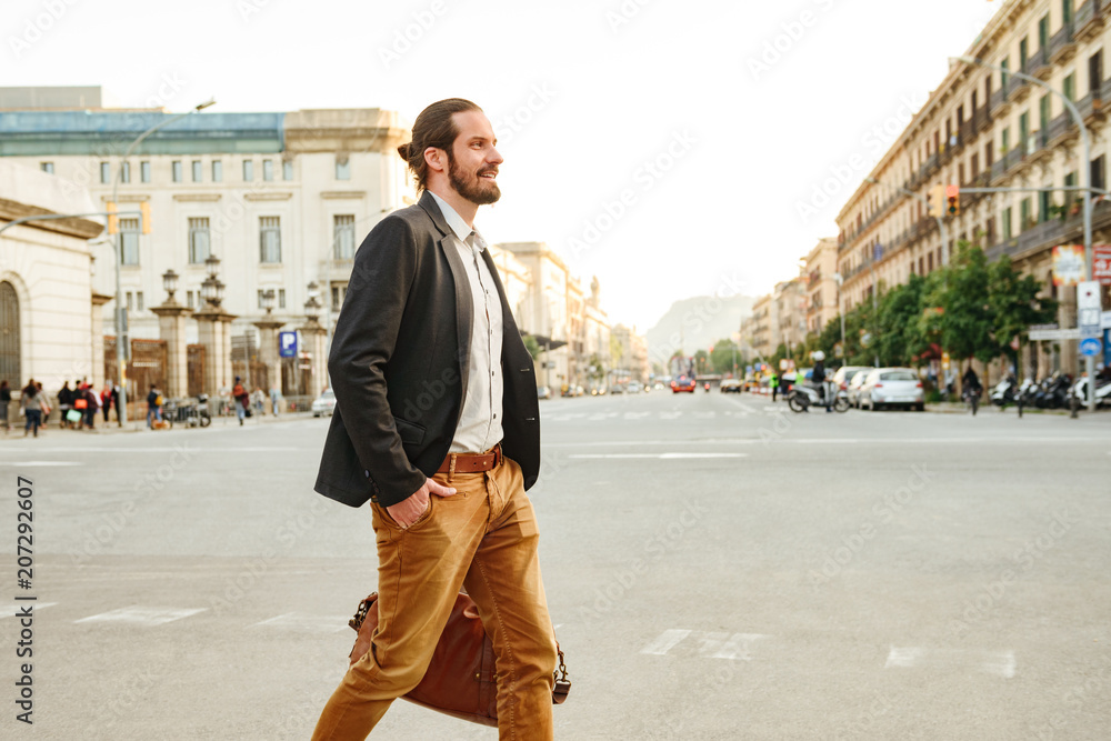 Portrait of confident successful man wearing stylish clothing walking across the road in city street, with leather male bag in hand