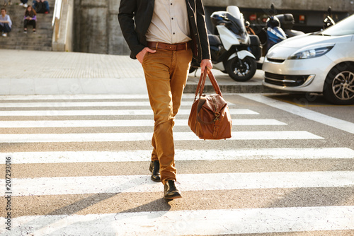 Cropped photo of stylish successful man in business wear walking through pedestrian crossing in downtown, with leather male bag in hand