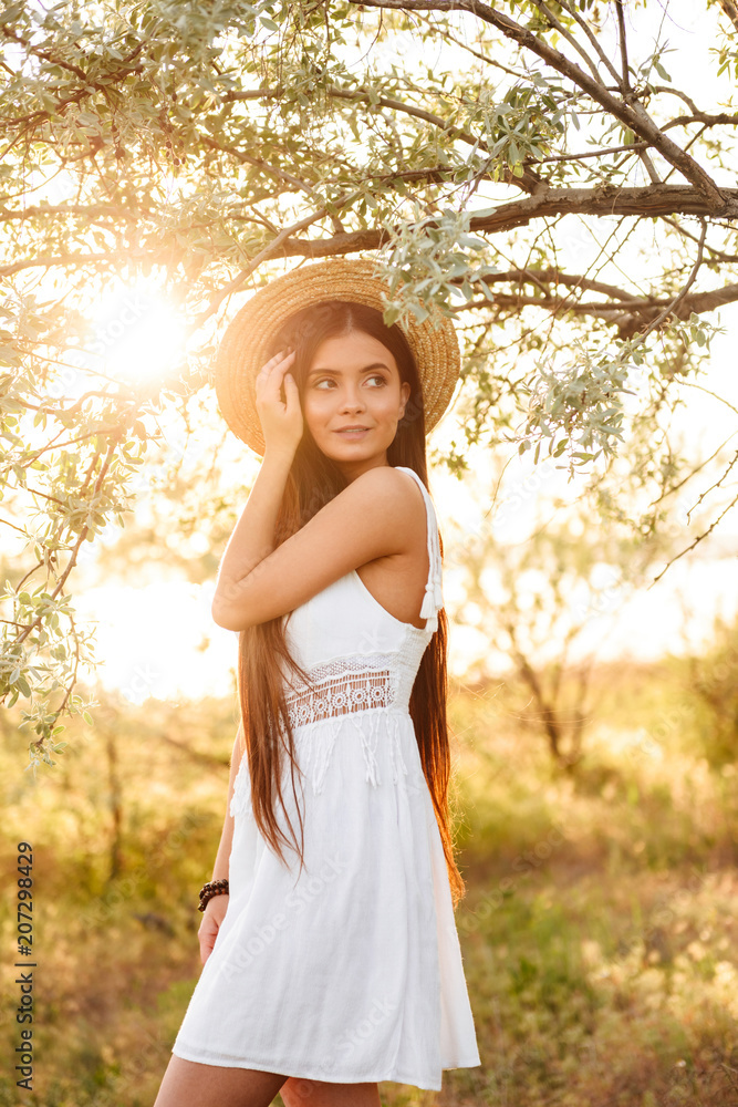 Photo of beautiful young woman with long dark hair wearing straw hat and white dress looking away, while standing under tree in nature during bright sunny day Photo of beautiful young woman with long dark hair wearing straw hat and white dress looking away, while standing under tree in nature during bright sunny day