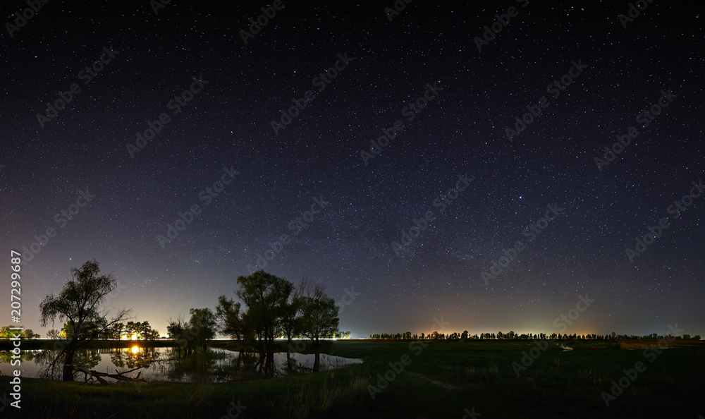 Space with the stars of the Milky Way in the night sky. The landscape with the river and trees is photographed on a long exposure.