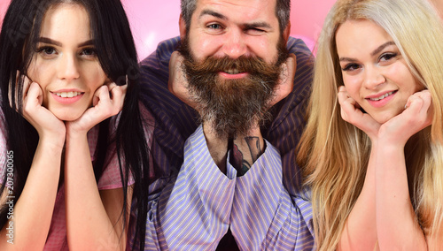 Man with beard and mustache, blonde and brunette girls. Girls with bearded macho, pink background. Best friends concept. Threesome on smiling faces lay near balloons, cheerful company, close up.