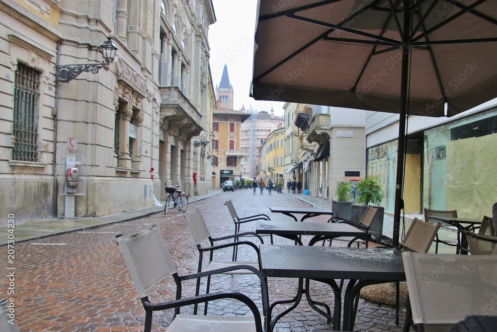 Naklejka premium Street cafe with chairs and tables in rainy weather. In the background the Parma Cathedral and Baptistery. Italy, South Europe.