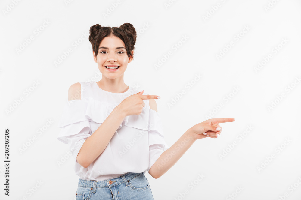 Portrait of european stylish woman with double buns hairstyle wearing braces on teeth pointing fingers aside at copyspace, isolated over white background