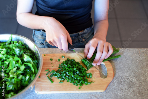 Teenage girl hands cutting fresh spring onion  with kitchen knife and wooden cutting board for preparing green salad for dinner on grunge grey table. Natural homemade cooking concept.