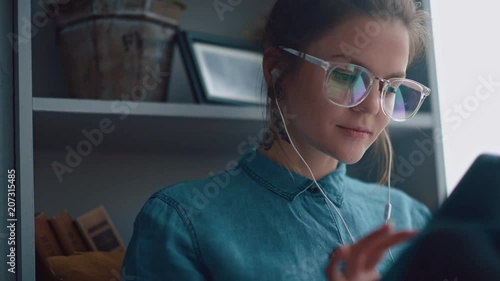 Wallpaper Mural Stylish student girl sits by the window (on windowsill) with a gadget, listens to music. She is wearing a denim shirt, screen of the tablet is reflected in lens of glasses. Calm girl reads an e-book Torontodigital.ca