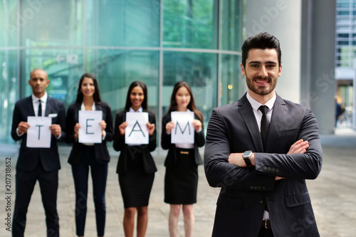 A group of businessman holds a sheet of paper with a letter that compose the word "TEAM" and in front of one of them he crosses his arms and smiles. Concept of: team work, finance, connection.