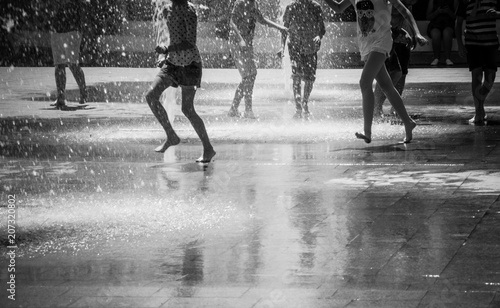 People walking among the place in summer with fountains and enjoying running in water with bare feet making the splashes. Black and white photo 
