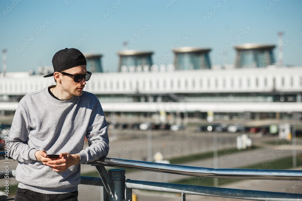 Handsome man traveler with phone near airport Pulkovo in Saint-Petersburg. He clothed in round sunglasses, black cap and grey sweater