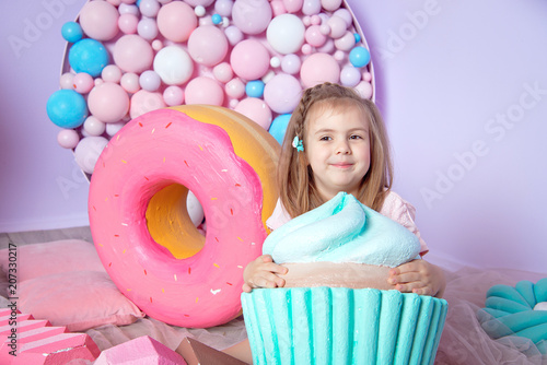 Little girl sitting in colorful room around big toyful candies, donut, lollipop. photo