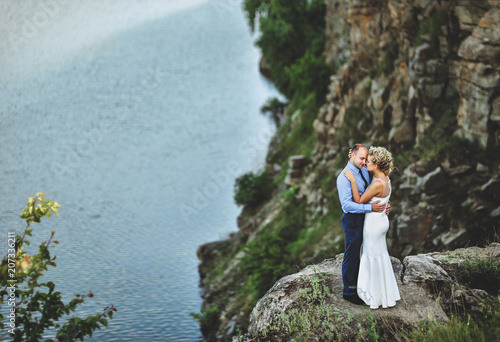 A nice bridegroom embraces a beautiful bride with curly hair and rests her forehead, standing on a precipice. Lovely newlyweds stand on a rock, against the background of rocks and the sea.