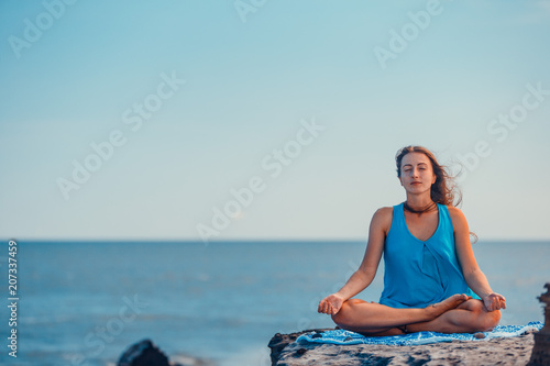 Young woman practices yoga and meditates in the lotus position on the rock at the sea beach.