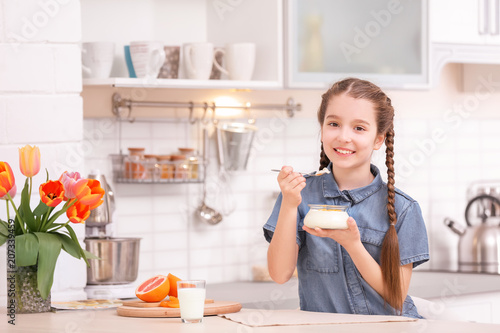 Cute girl eating tasty yogurt at table in kitchen