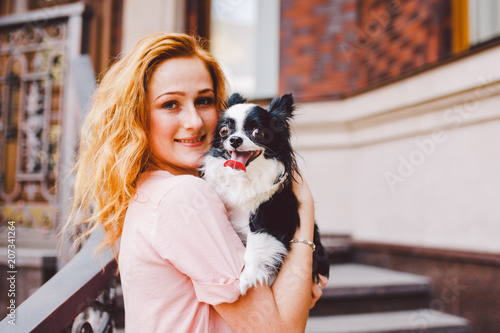 A beautiful young woman with red long hair is holding a small, cute funny big-eyed dog of two flowers, a black-and-white pet of the breed of hichuahua against a house of red brick in summer © Elizaveta
