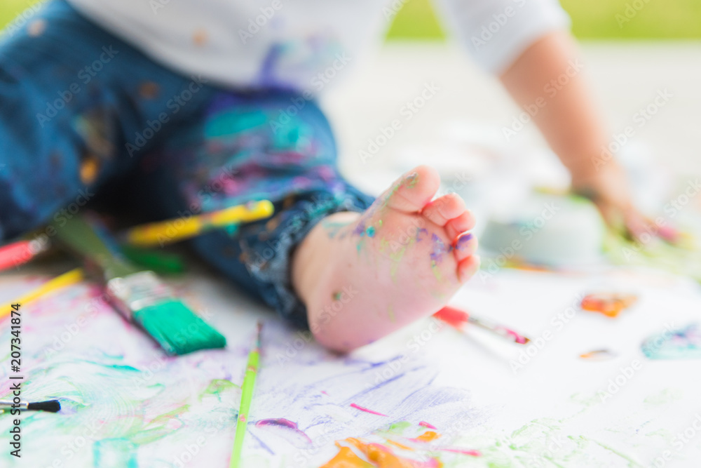 White toddler foot with splattered paint surrounded by paint brushes and paint covered paper. White toddler foot with splattered paint surrounded by paint brushes and paint covered paper.