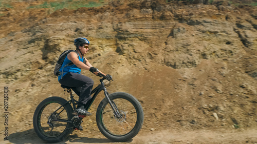 Fat bike also called fatbike or fat-tire bike in summer driving on the road. The guy rides by the hill on a sand clay path, behind him the shore by the sea.