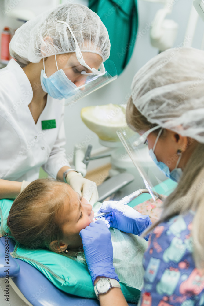 Child with a mother at a dentist's reception. The girl lies in the chair, behind her mother. The doctor works with an assistant. Procedure for drilling a tooth. Setting a temporary seal. Using the x Child with a mother at a dentist's reception. The girl lies in the chair, behind her mother. The doctor works with an assistant. Procedure for drilling a tooth. Setting a temporary seal. Using the x