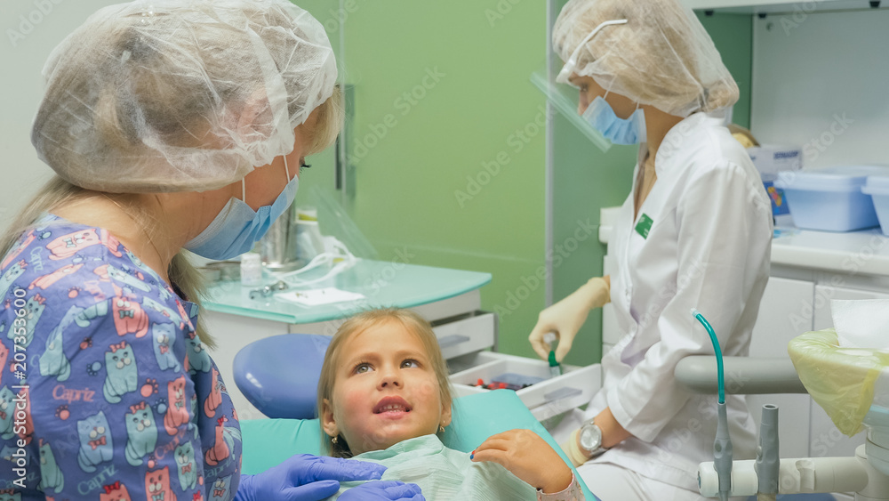 Child with a mother at a dentist's reception. The girl lies in the chair, behind her mother. The doctor works with an assistant. Procedure for drilling a tooth. Setting a temporary seal. Using the x Child with a mother at a dentist's reception. The girl lies in the chair, behind her mother. The doctor works with an assistant. Procedure for drilling a tooth. Setting a temporary seal. Using the x
