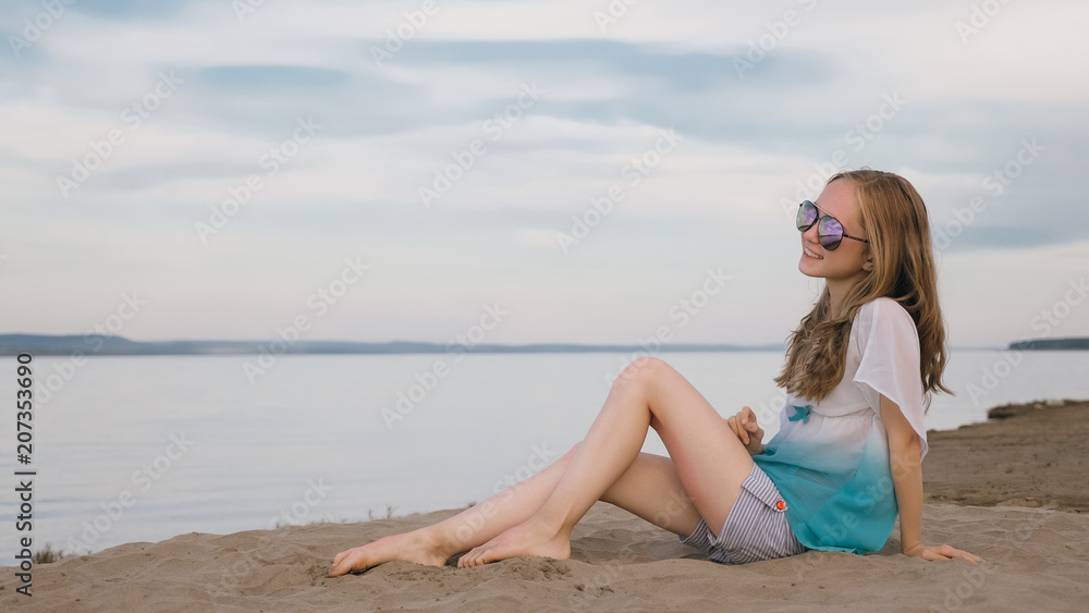 One beautiful teenage girl with brown hair outside on a beautiful summer day. The woman is sitting on the beach. The sun was gone in the sunset. The girl is genuinely genuinely genuinely smiling and