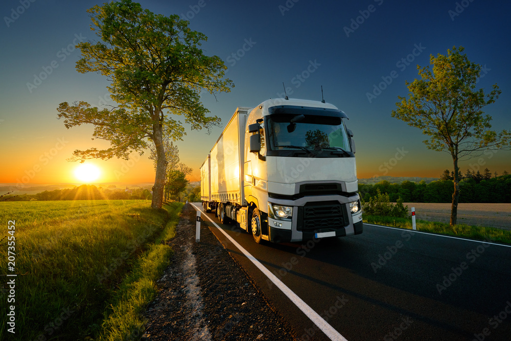 White truck driving on the asphalt road between trees in a rural landscape at sunset