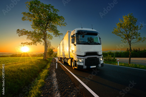 White truck driving on the asphalt road between trees in a rural landscape at sunset