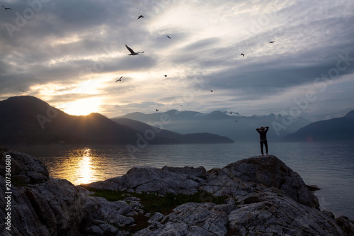 Woman enjoying the beautiful Canadian Mountain Landscape during a vibrant sunset. Taken on a rocky Island in Howe Sound near Vancouver, BC, Canada.