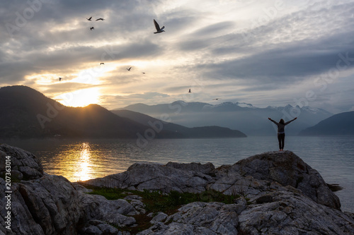 Woman with open arms is enjoying the beautiful Canadian Mountain Landscape during a vibrant sunset. Taken on a rocky Island in Howe Sound near Vancouver, BC, Canada.