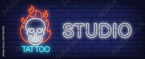 Tattoo studio neon sign. Smiling scull on fire and big inscription on dark brick wall. Vector illustration in neon style for tattoo salon or luminous advertisement